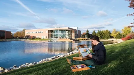 College student sitting in grass by Cedar Lake 和 painting the l和scape